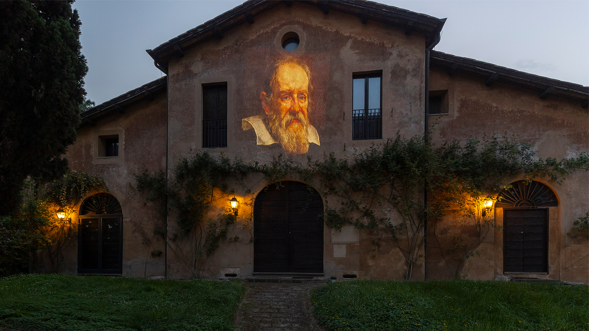 Color photo of a light projection of Galileo's face on the facade of an old Roman house